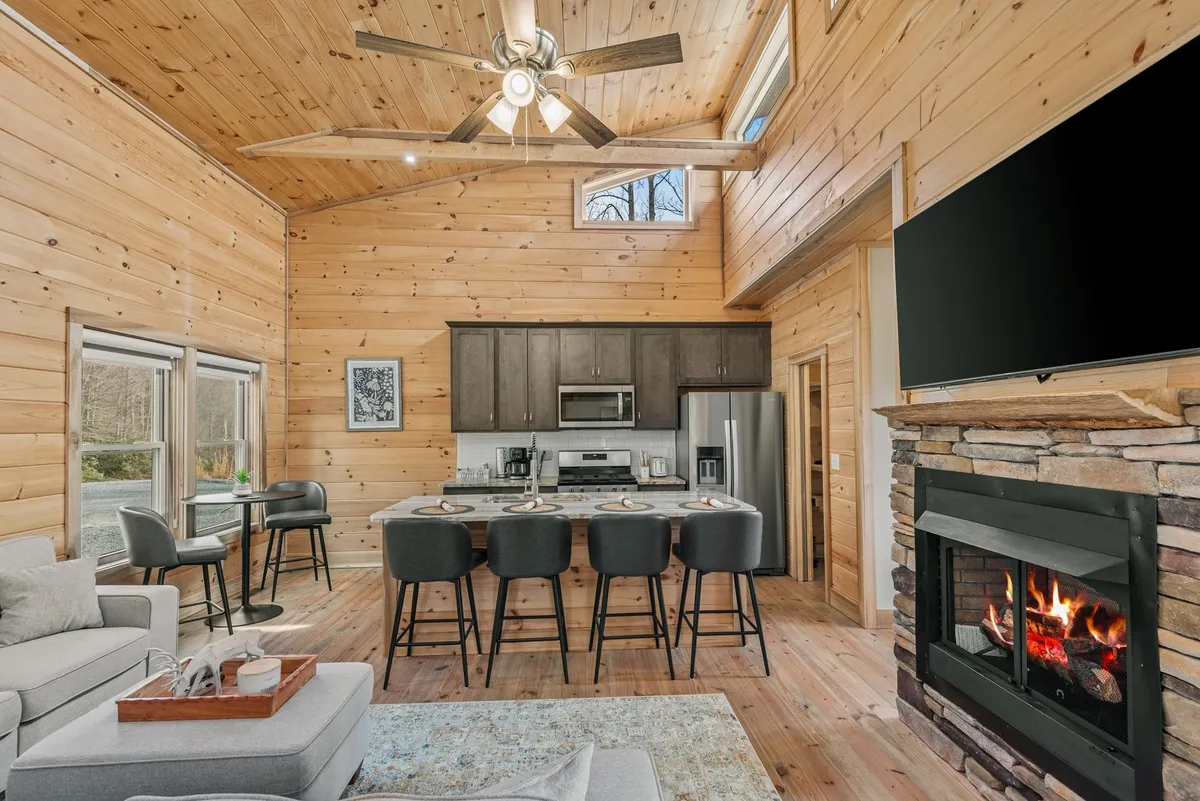 A view that unifies the kitchen island, comfortable sofa, windows, and a glowing fireplace, all framed by natural wood elements. The scene radiates warmth and the sense of a central hub for cooking, relaxing, and gathering.