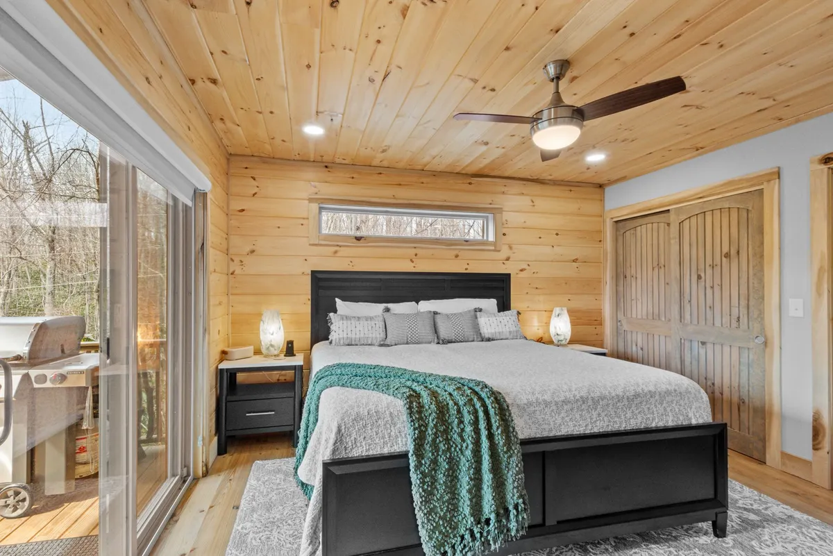 A spacious king bedroom with a large bed dressed in decorative pillows, warm wooden paneling, and a sliding glass door that opens to the outdoors. Nightstands with soft-glowing lamps add to the relaxing, inviting ambiance.
