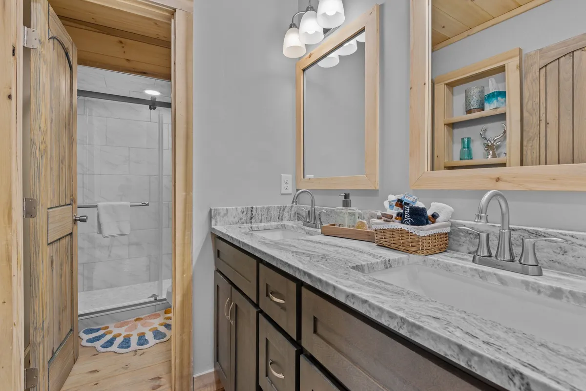 A modern ensuite bathroom with double sinks set in a granite countertop, wooden cabinetry, and a glass-enclosed shower. The space is bright, organized, and conveys a sense of luxury and relaxation.