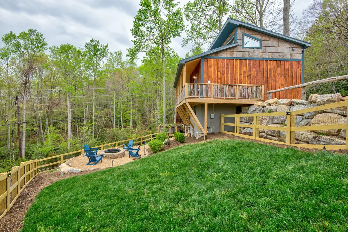 A welcoming exterior shot of a modern wooden house nestled among trees, featuring a cozy fire pit with seating. The scene is tranquil, with green grass and a relaxing seating area perfect for gatherings or quiet contemplation after a day in the mountains.