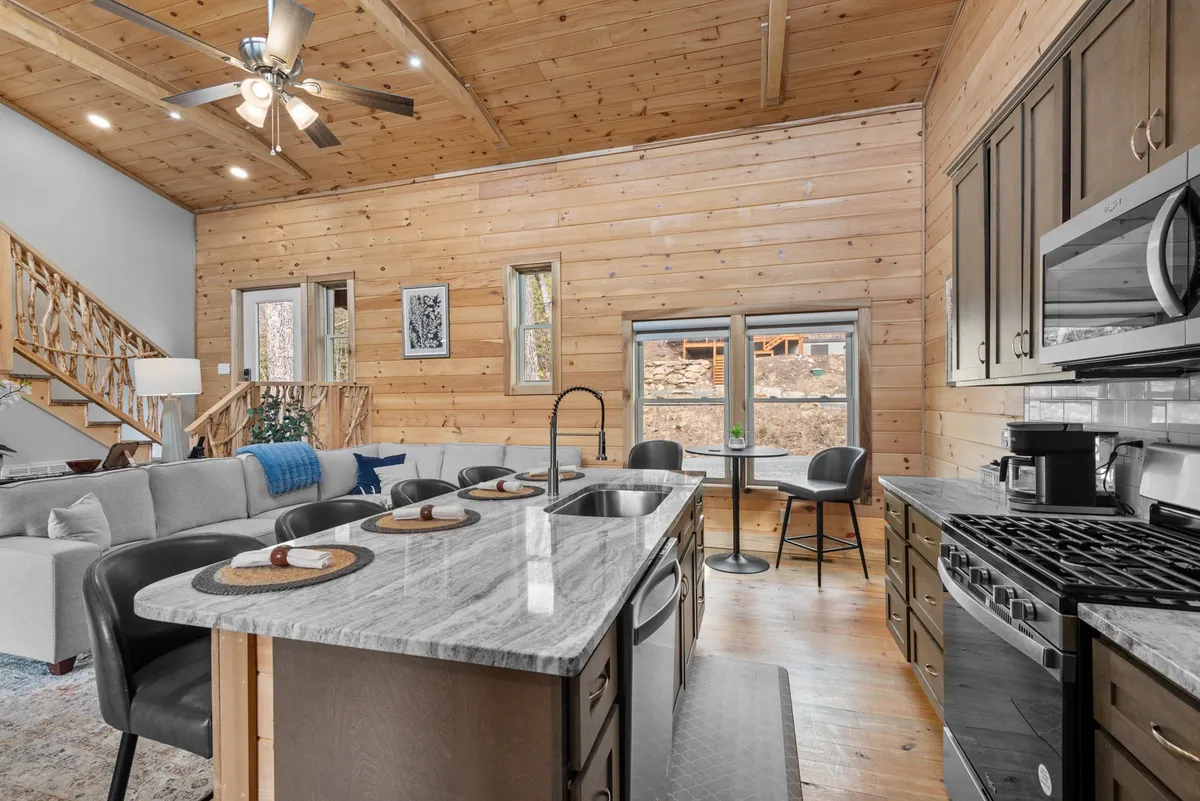 A bright, airy interior showcases a kitchen island with a gleaming marble countertop, a comfortable sofa, large windows streaming natural light, and a sturdy wooden staircase. The interplay of light and space evokes warmth and luxury.