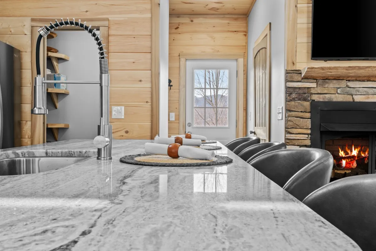 The focus is on a modern granite kitchen countertop with stylish stools, a sleek faucet, and the cozy backdrop of a fireplace. The arrangement suggests a space perfect for both entertaining and intimate gatherings.