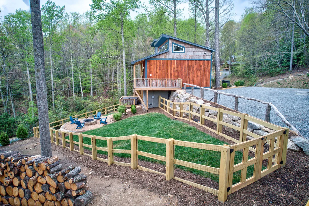 The photo shows the modern wooden house with a fenced yard, a welcoming fire pit area, and surrounding trees, along with stacked logs nearby. The setting is both peaceful and playful, offering ample space for families and pets to explore and enjoy the outdoors.