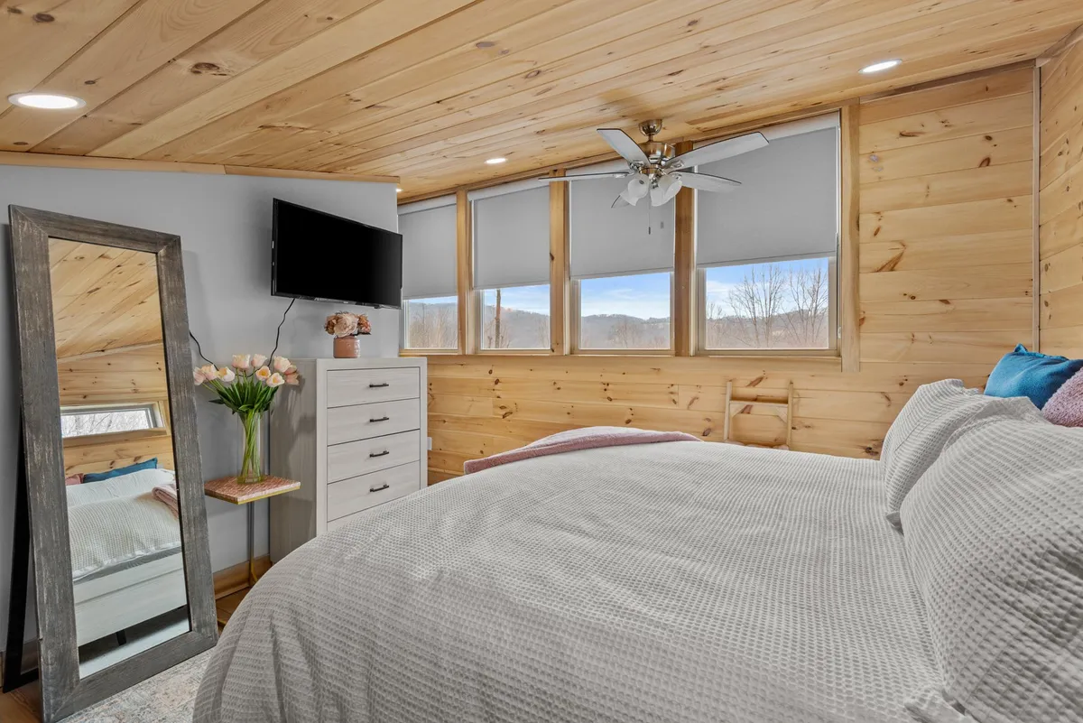 A cozy bedroom featuring a bed with textured bedding, a large mirror, flower vase, wall-mounted television, and generous windows revealing scenic mountain views. The room feels peaceful and well-appointed, blending comfort with modern amenities.
