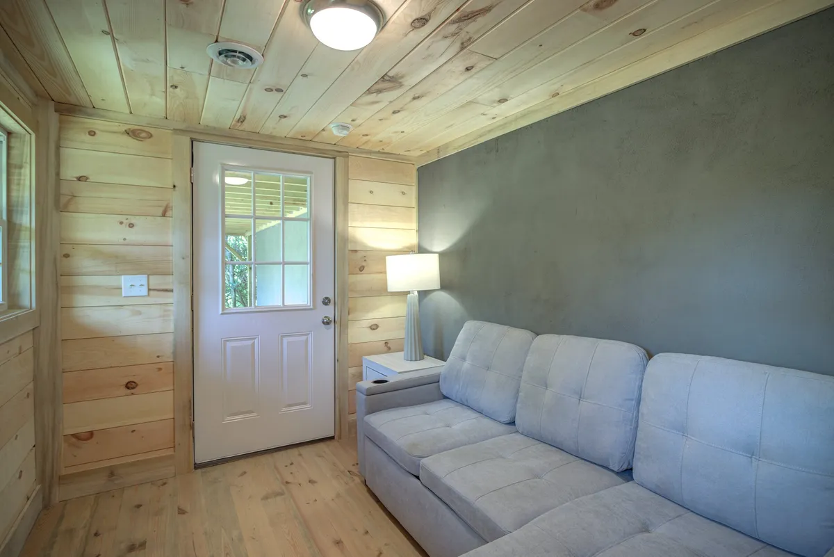 The image shows a serene, softly lit room with a sofa, a door, a warm lamp, and wood-paneled walls. The inviting atmosphere highlights the space’s adaptability for reading, relaxing, or socializing.