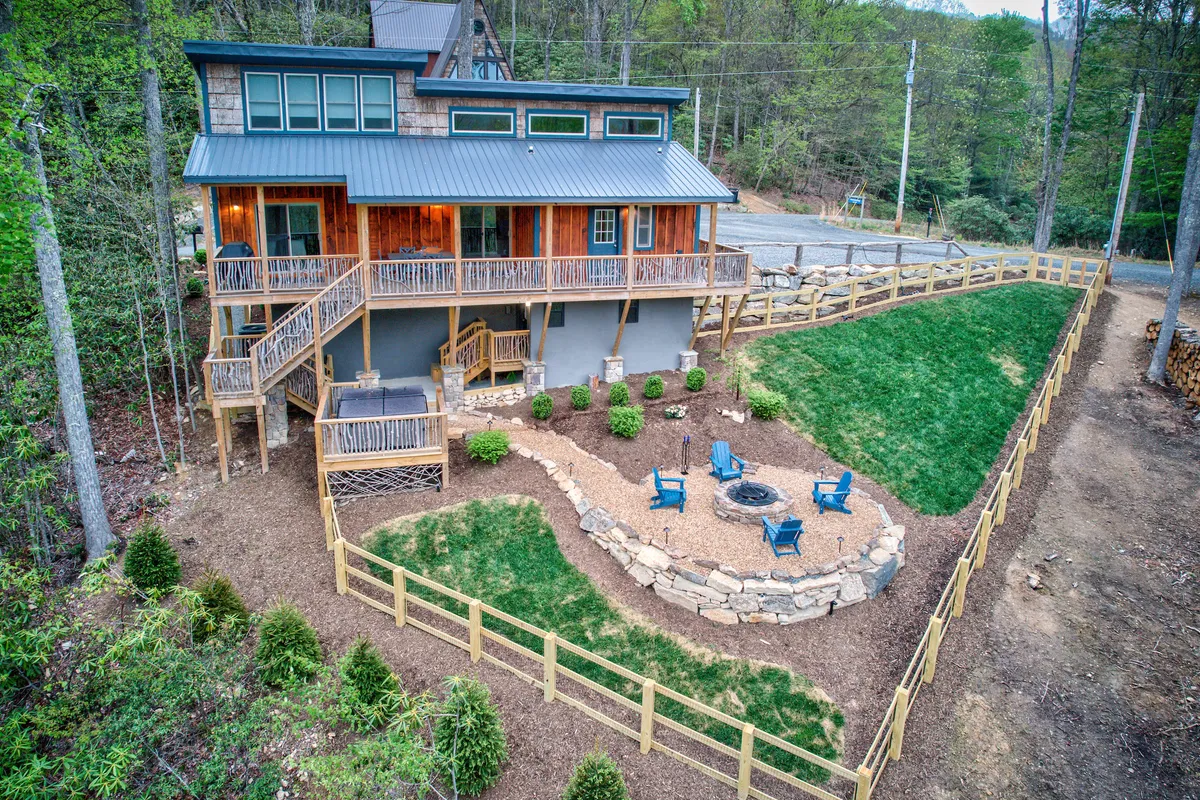 The image showcases the exterior of a charming mountain house set among lush landscaping, featuring a spacious wooden deck outfitted with stylish blue chairs and a modern fire pit. The setting is ideal for gatherings, with inviting spaces for relaxing, sharing stories, and enjoying the natural beauty of Beech Mountain.