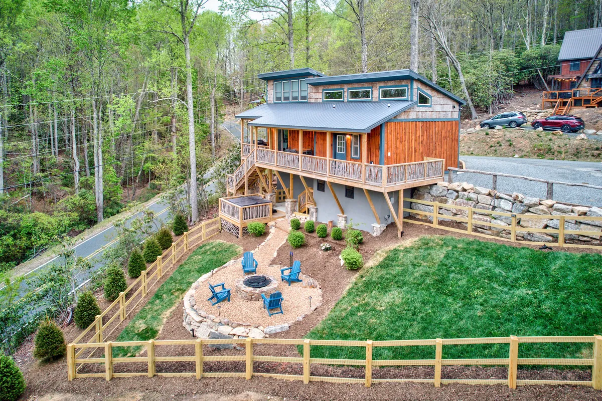 The inviting wooden house is featured with a circular fire pit area surrounded by blue chairs, set amidst lush greenery. The setting exudes a sense of relaxation and communal gathering after a day outdoors.
