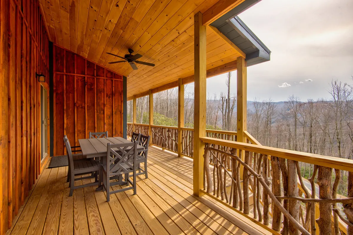 The photo features a spacious wooden deck with a dining table and chairs, bordered by a wooden railing. In the distance, stunning mountain views create a breathtaking backdrop, while the deck’s setup hints at outdoor dining and relaxation.